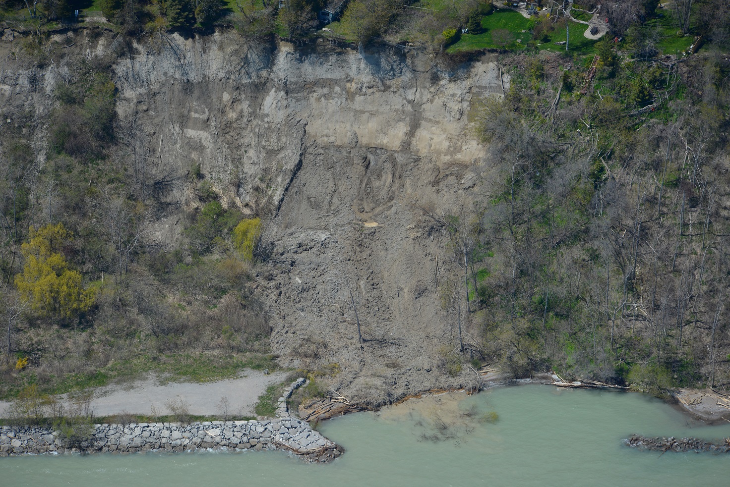This image was taken from a helicopter and shows a landslide that has settled near the base of a very tall section of the Scarborough Bluffs. At the bottom of the photo, Lake Ontario is visibly turbid due to soil from the landslide being washed into the water. At the bottom left of the image is an dirt access road behind a shoreline revetment structure constructed of large 5 tonne armourstone blocks. The face of the bluff on either side of the slope failure is vegetated with shrubs and trees.