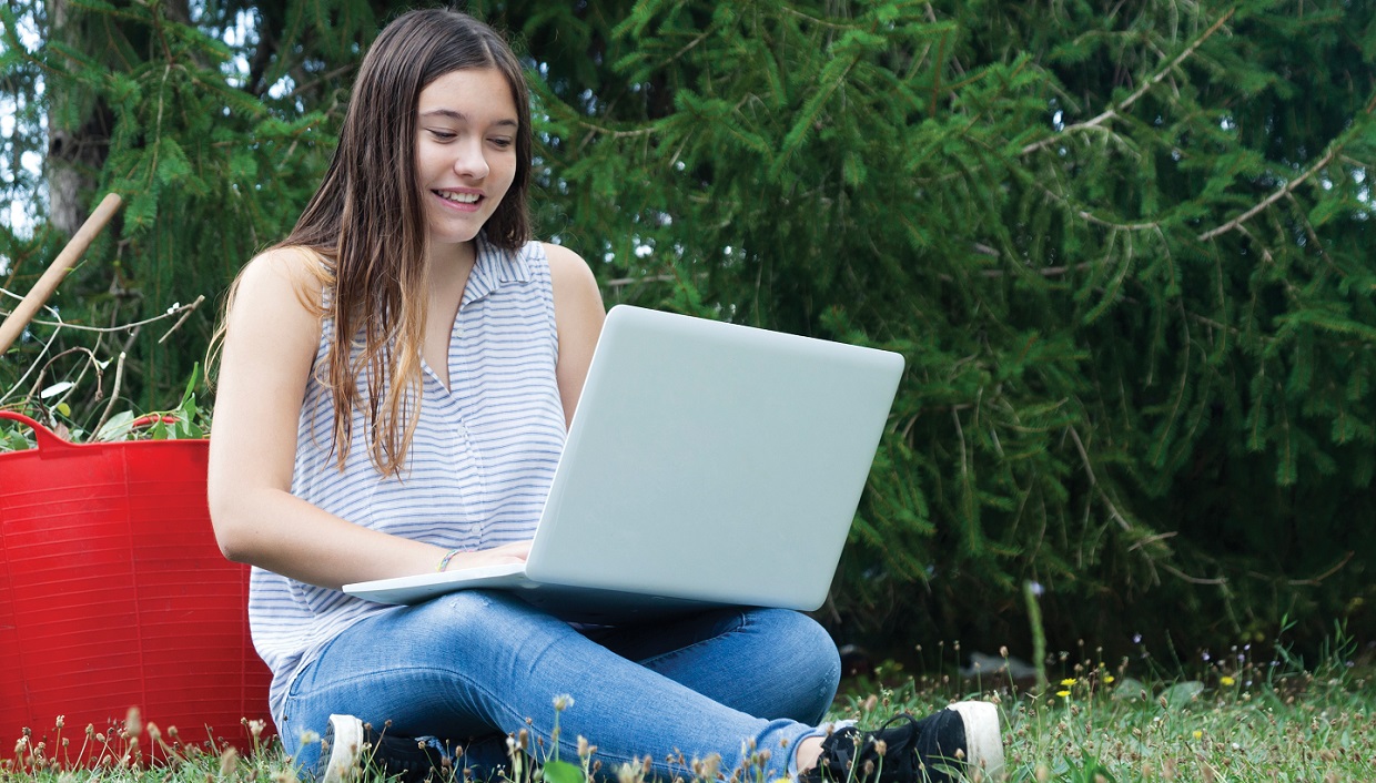 female high school student works on laptop while sitting on grass in park